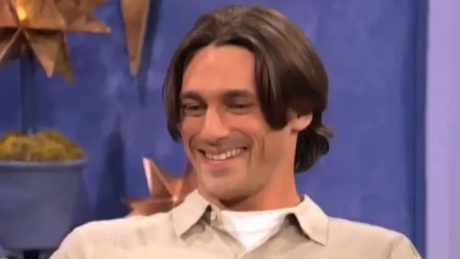 A man with shoulder-length brown hair and a beige shirt smiles while sitting indoors, with blue walls and decorative items in the background.