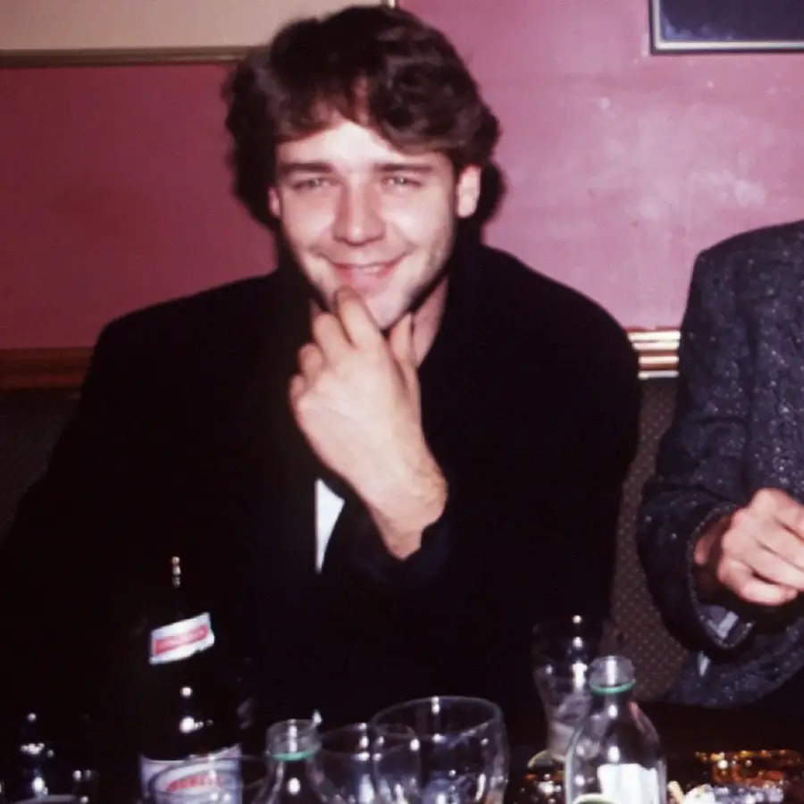 A young man with brown hair smiles and touches his chin while sitting at a table with empty glasses, bottles, and someone partially visible beside him against a maroon wall.