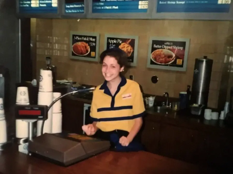 A young woman in a yellow and blue uniform smiles behind a fast-food counter, with a register, cups, and menu signs featuring apple pie and shrimp on the wall behind her.