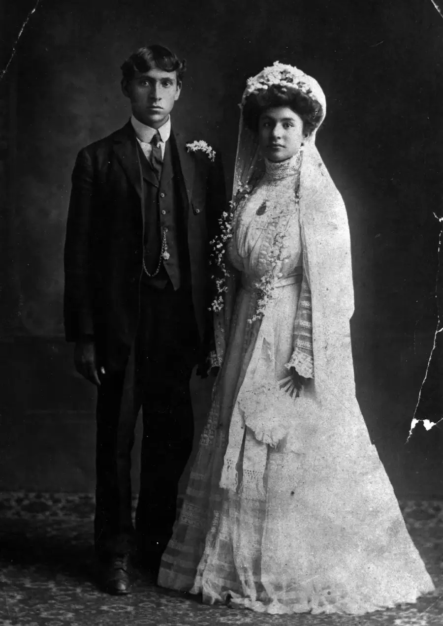 A black-and-white photo of a bride and groom in formal wedding attire. The groom wears a suit with a boutonniere, while the bride wears a long white dress, veil, and floral headpiece, standing side by side.