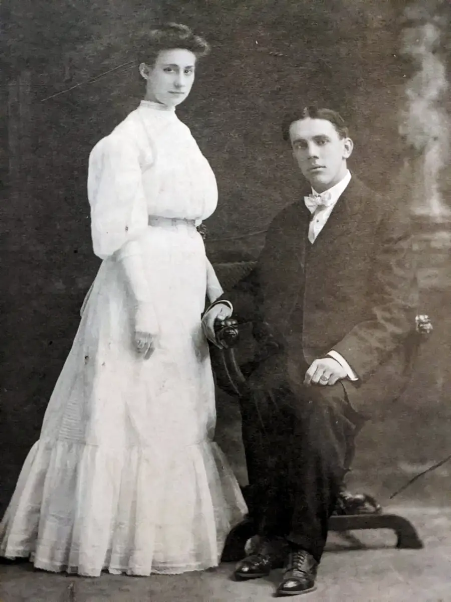 A black-and-white photo of a woman in a long white dress standing next to a seated man in a dark suit and bow tie, both looking at the camera, in a vintage portrait setting.