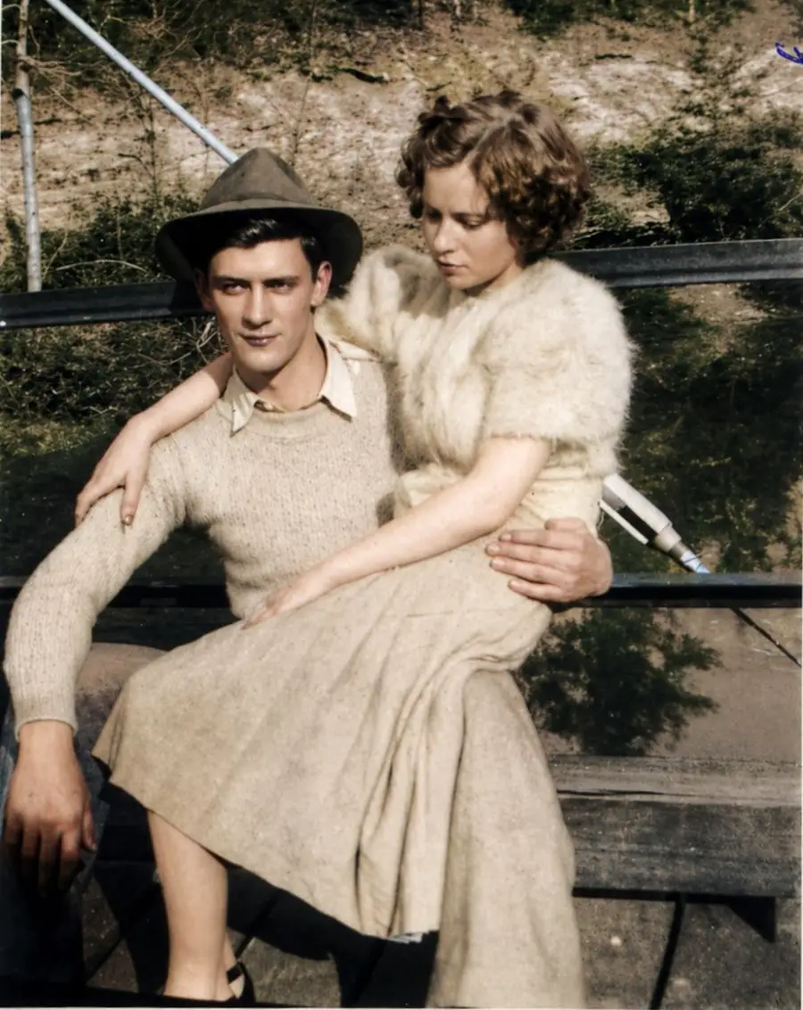 A young man in a hat and sweater sits on a bench holding a woman in a fur-collared dress on his lap. They are outdoors, with foliage in the background. Both appear relaxed and close.