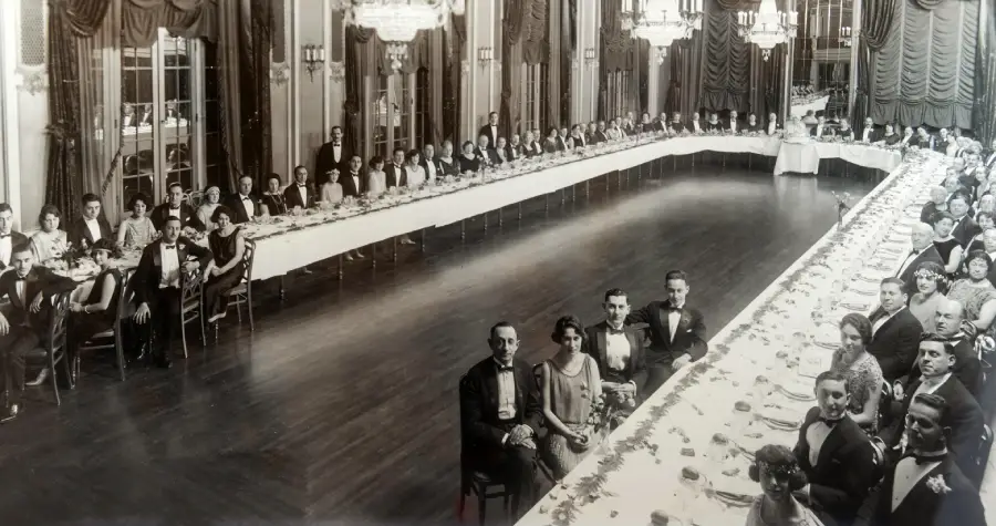 A large group of people in formal attire are seated around a long, U-shaped banquet table in an ornate, elegant ballroom with chandeliers and tall windows.