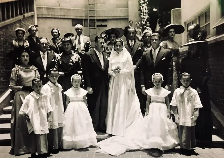 A black-and-white photo of a wedding party outdoors, featuring a bride and groom at the center, surrounded by bridesmaids, groomsmen, flower girls, and other guests, all in formal attire, posing on steps.