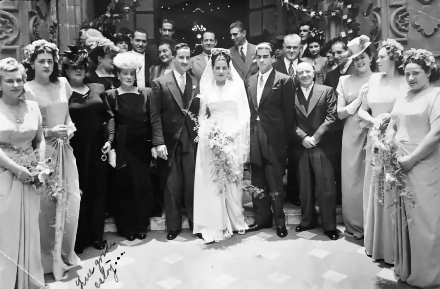 A black-and-white photo of a wedding party, with the bride and groom at the center, surrounded by family and friends dressed in formal attire, standing outside a decorated building.