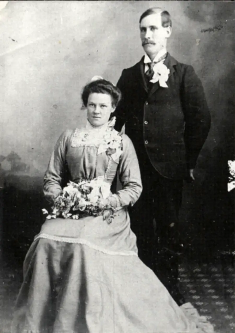 A vintage black-and-white photo of a woman in a long dress seated with flowers, and a man in a suit and bow tie standing beside her, possibly taken at a formal event or wedding.