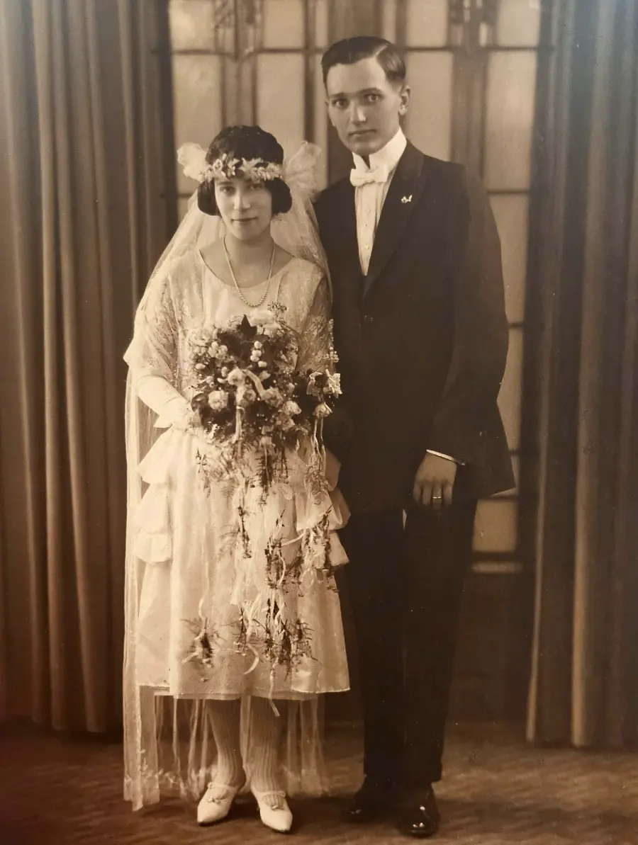 A bride in a vintage dress and veil stands beside a groom in a dark suit and bow tie. They pose together indoors, with a bouquet and curtains behind them, in a sepia-toned wedding portrait.