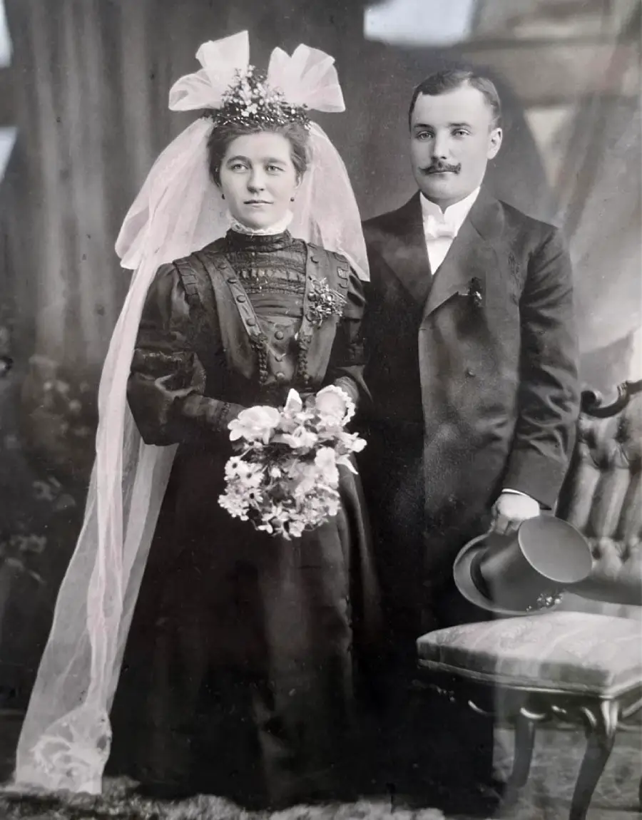 Old black-and-white photo of a bride in a long dress, veil, and flower crown holding a bouquet, standing next to a groom in a suit holding a top hat, posing for a formal wedding portrait.