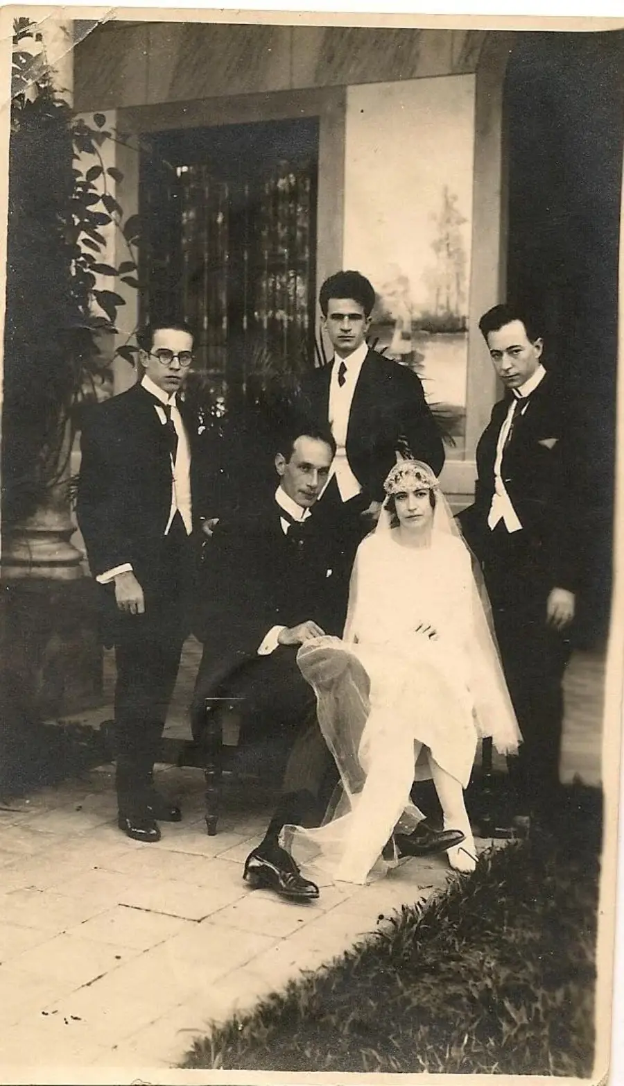 A vintage black-and-white photo of a bride in a veil and gown seated next to a man in a suit, with three other men in suits standing around them outdoors.