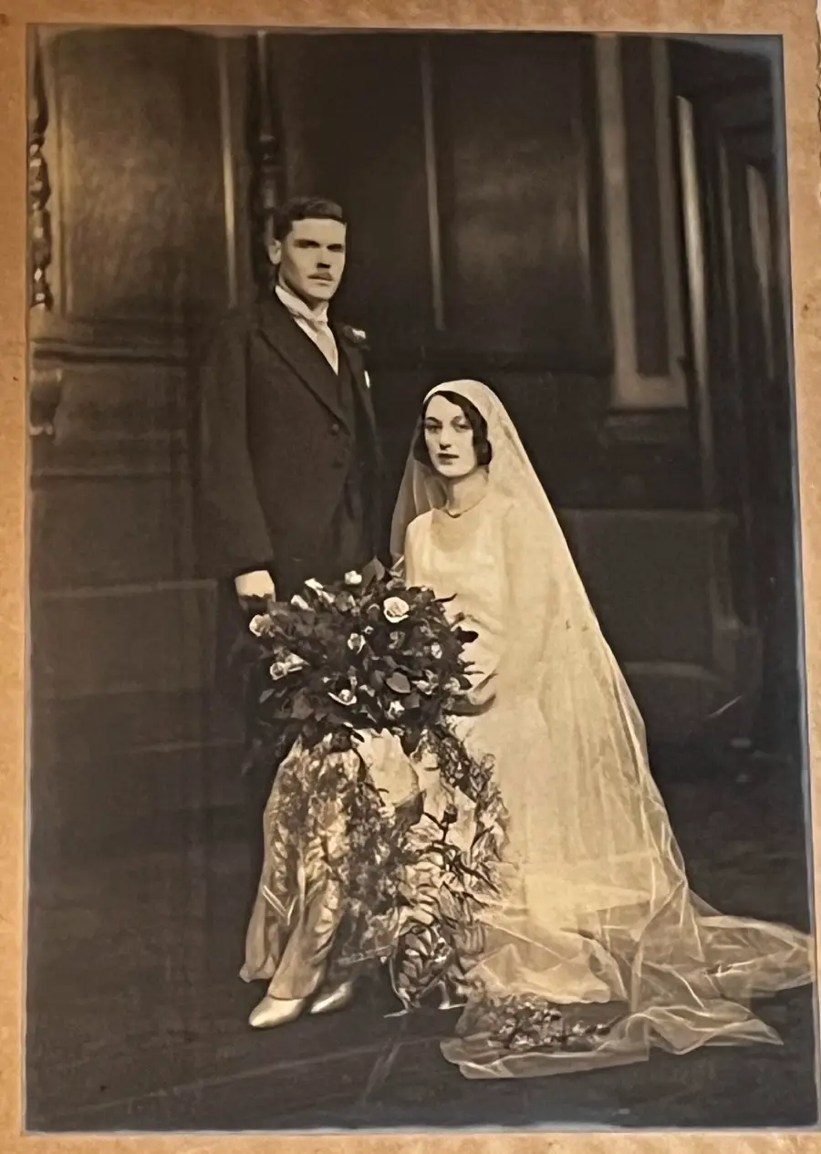 A vintage black-and-white photo of a bride in a long veil and a groom in a suit posing together indoors. The bride is seated, holding a large bouquet, while the groom stands beside her.