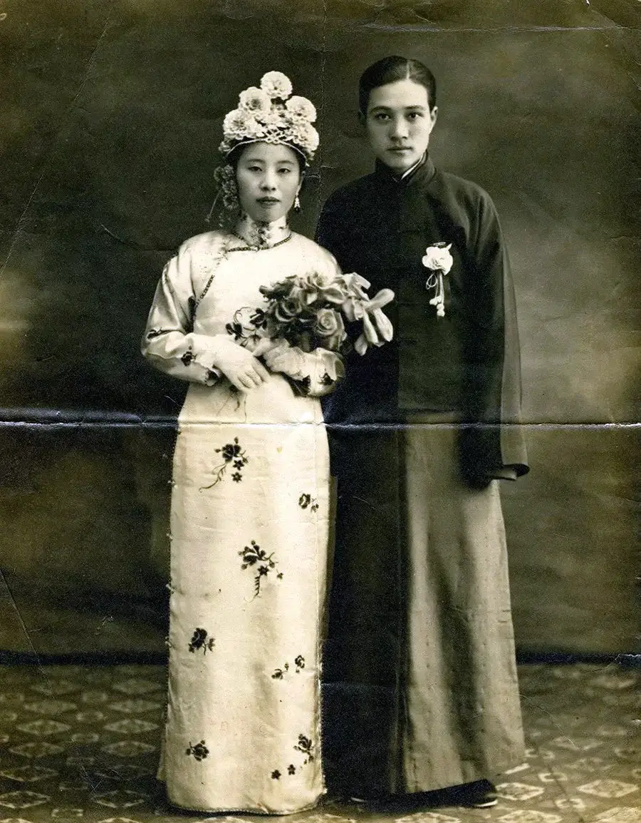 Two people in traditional attire pose for a formal portrait. The person on the left wears an embroidered dress, elaborate headpiece, and holds flowers. The person on the right wears a long dark robe with a floral pin.