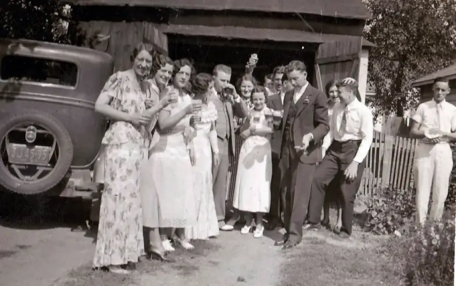 A group of people in vintage clothing smile and pose together outdoors near a car and wooden building, appearing to celebrate or attend a gathering on a sunny day.