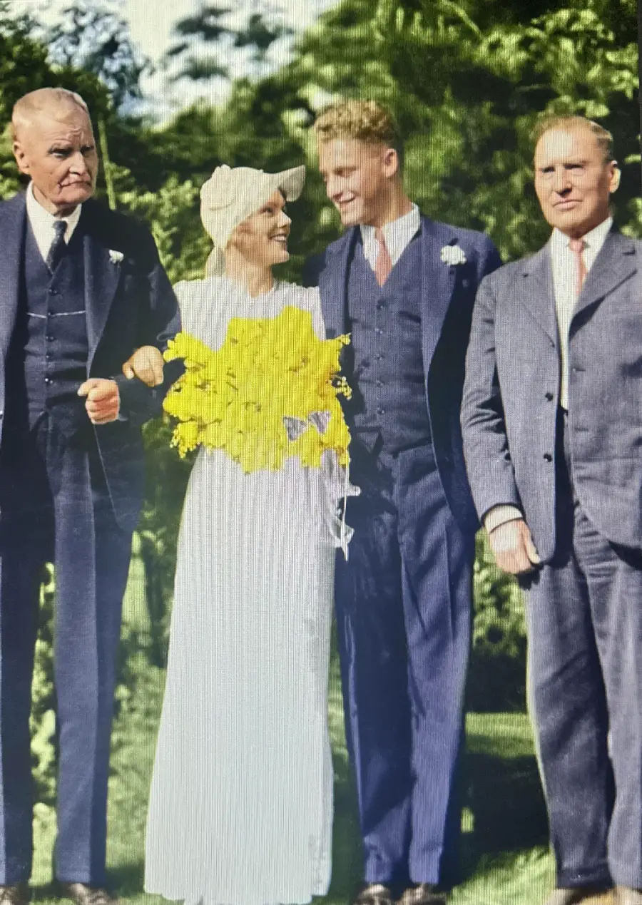 A bride holding a yellow bouquet stands outside with three men in suits. The group poses for a photo, smiling, with greenery in the background.