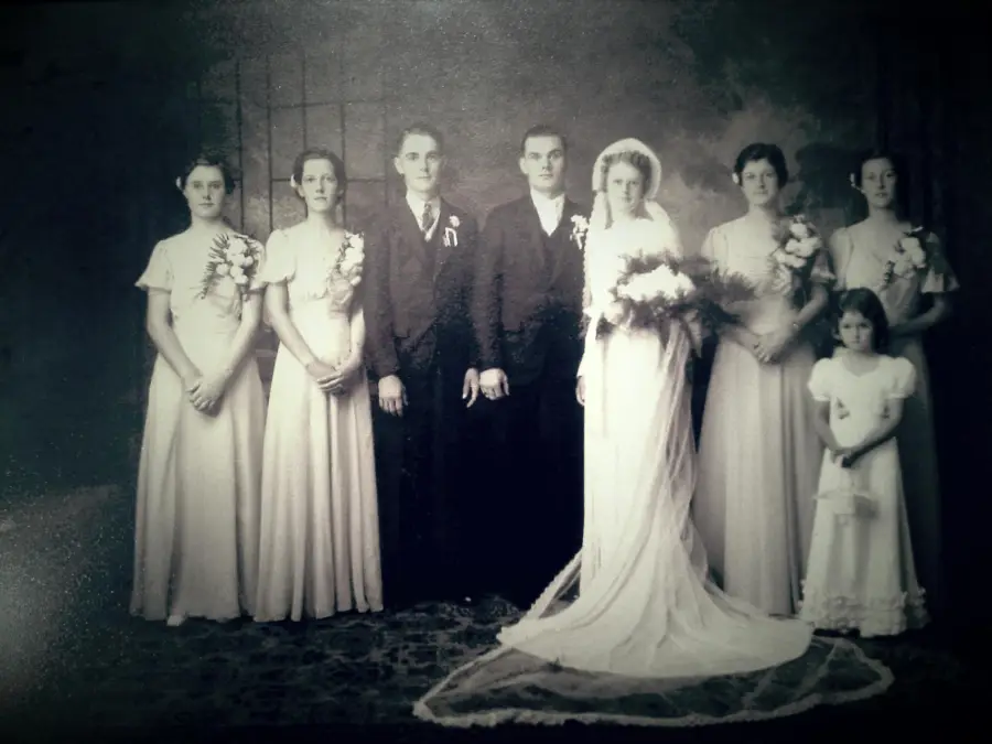 A vintage black-and-white wedding photo shows a bride, groom, bridesmaids, groomsman, and a young girl in formal attire, standing in a studio setting with neutral expressions.