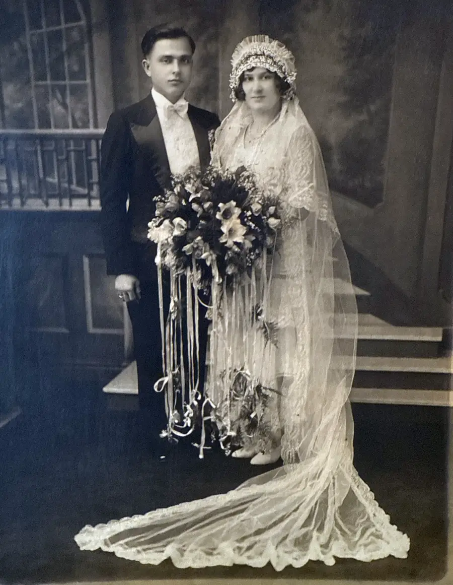 A black-and-white photo of a bride in a long lace gown and veil holding a large bouquet, standing beside a groom in a tuxedo, posed in a vintage studio with a painted backdrop and steps.