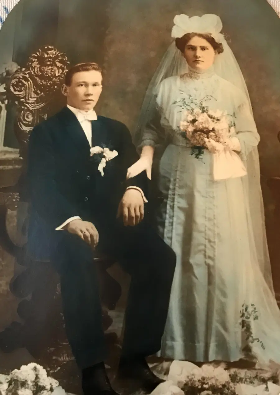 A vintage sepia-toned photo of a bride in a long dress and veil holding flowers, standing beside a seated groom in a dark suit, possibly from the early 1900s.