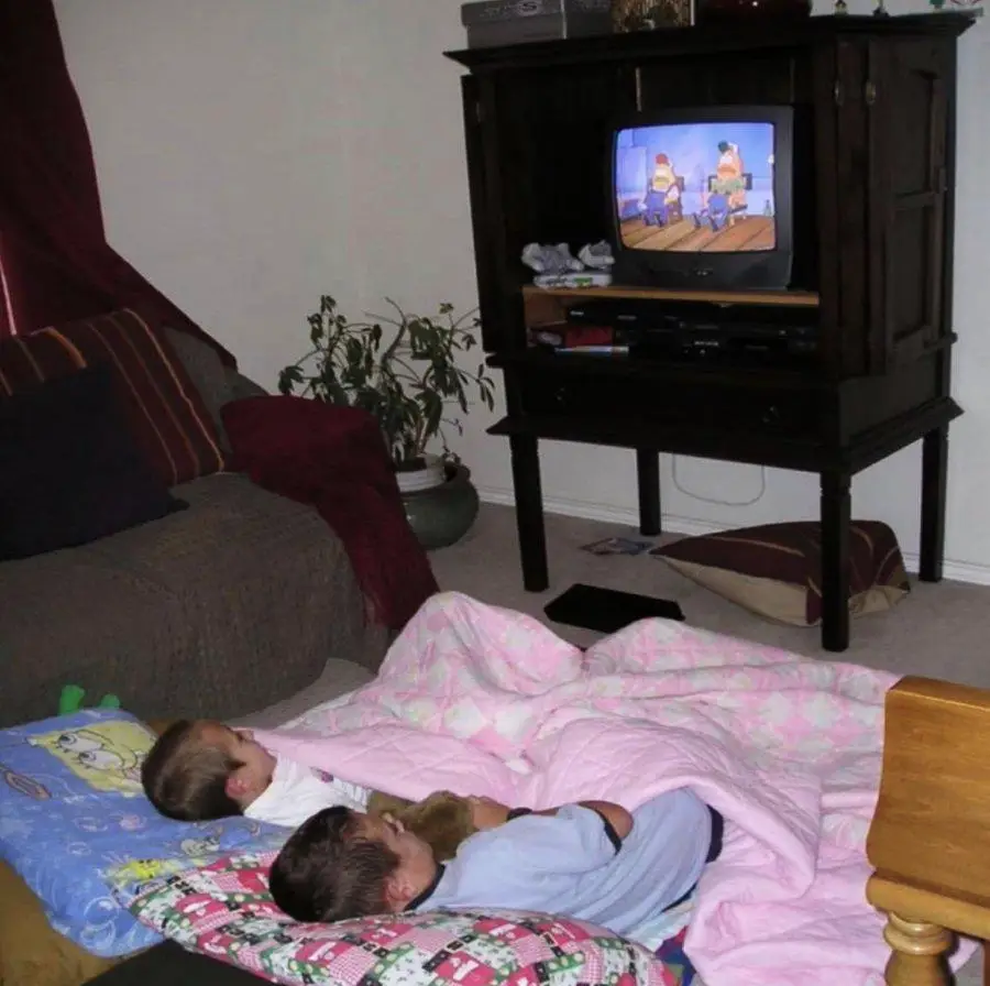 Two kids lie on the floor under blankets, watching a cartoon on an old TV. A couch, plant, and dark wooden TV stand are visible in the cozy living room.