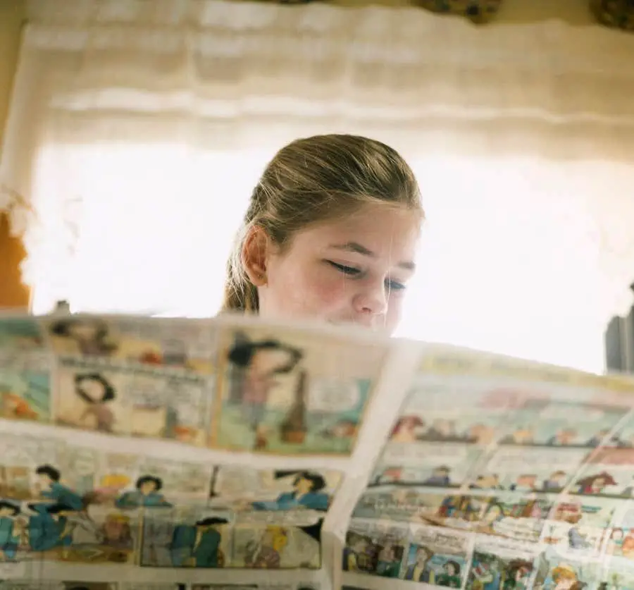 A young girl with light brown hair reads a newspaper comic section indoors, with sunlight streaming through a window behind her.