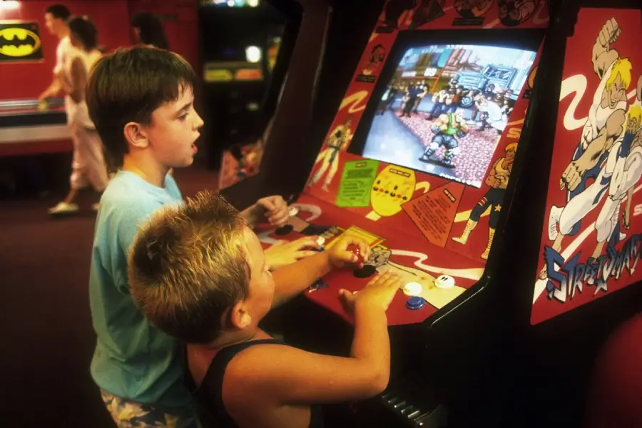 Two boys play a Street Fighter arcade game in an arcade. The older boy uses the controls while the younger boy watches. Colorful artwork and other arcade machines are visible in the background.