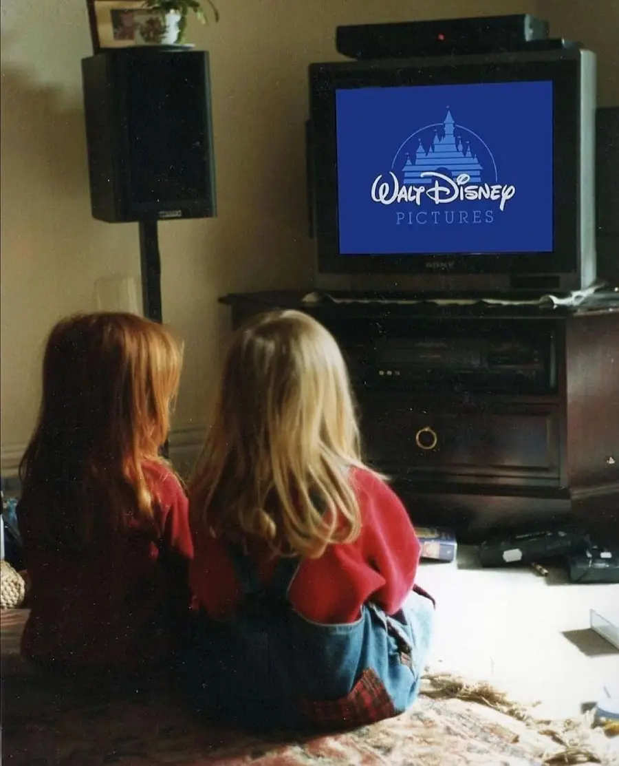 Two young children sit on the floor watching a TV showing the Walt Disney Pictures logo. The room has a TV stand, speakers, and scattered objects.