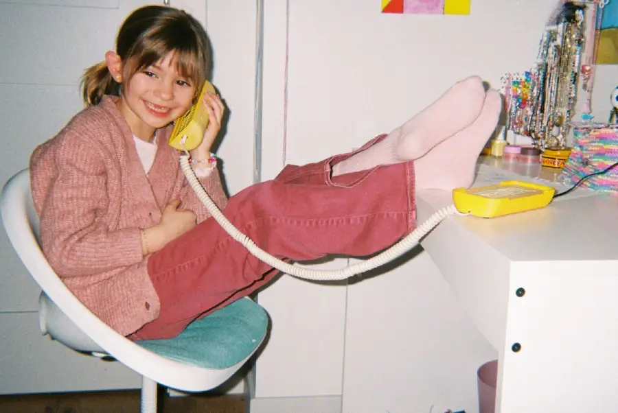 A smiling child sits in a chair with feet up on a desk, talking on a yellow corded phone. She wears pink socks, red pants, and a pink sweater. Jewelry and colorful items are on the desk.