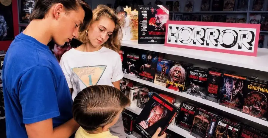 Three kids stand by a display of horror VHS tapes in a video store. One holds a tape while the others look on. A white sign with "HORROR" in black letters is above the shelves filled with scary movie covers.