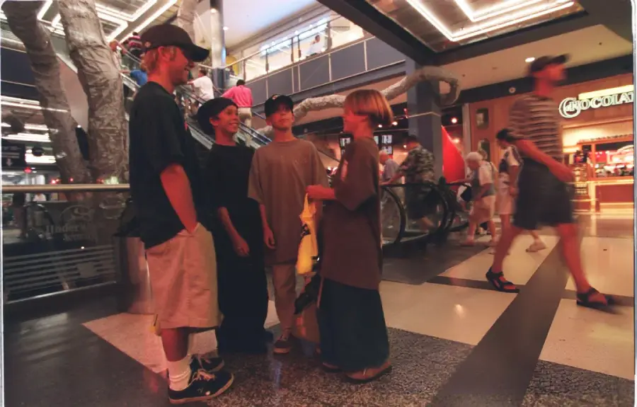 Four young people stand together talking inside a shopping mall near an escalator. Other shoppers walk by, and a store called “Chocolateria” is visible in the background. The scene is casual and lively.