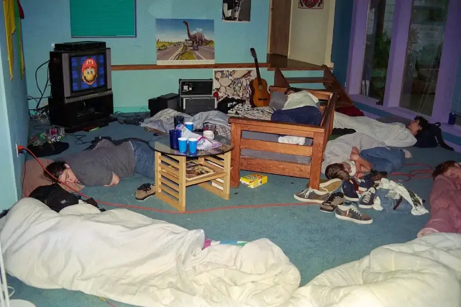 A messy living room with several people sleeping on the floor in blankets, a TV on, scattered shoes, cups on a small table, a guitar, and posters on the wall.
