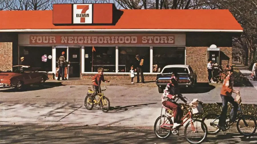 Children ride bikes outside a vintage 7-Eleven store with a sign reading "Your Neighborhood Store." Several cars are parked in front, and people are near the entrance under a bright red roof. Trees are visible in the background.
