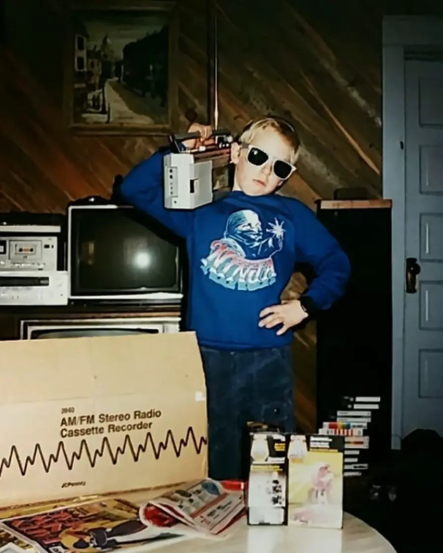 A young boy in sunglasses poses with a boombox on his shoulder, standing next to a stack of electronics and cassette tapes. There’s an open box labeled "AM/FM Stereo Radio Cassette Recorder" in front of him.