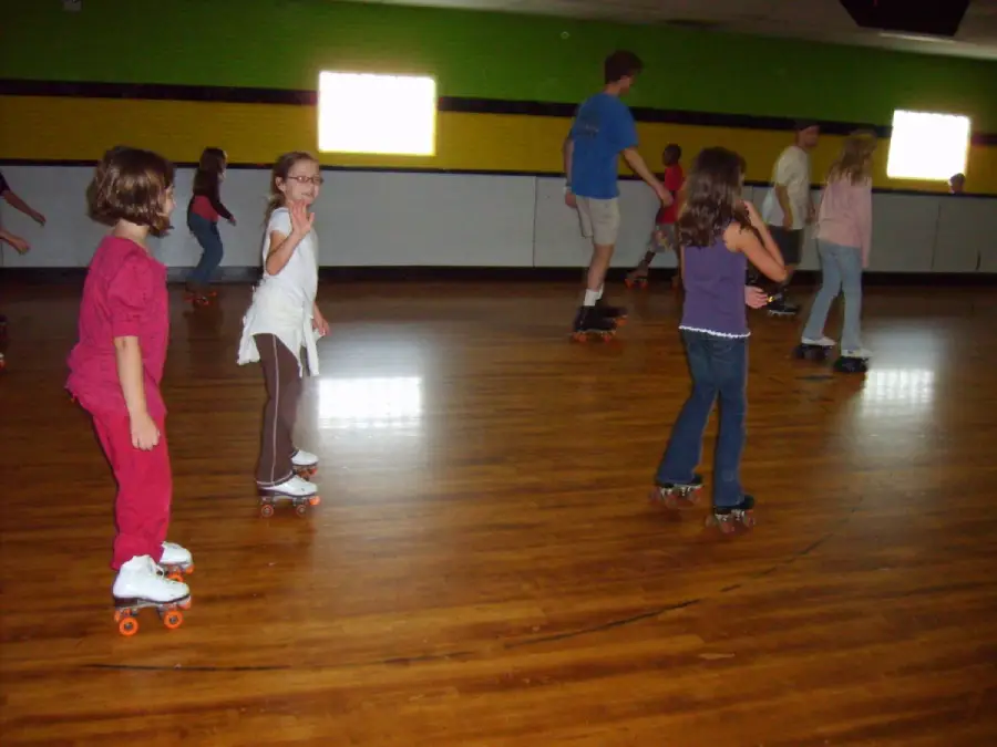 Children and adults roller skating on a shiny wooden floor in an indoor rink with green and yellow walls and bright windows in the background.