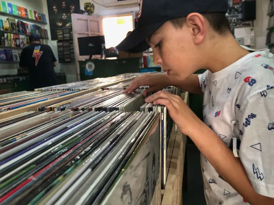 A young boy wearing a cap browses through vinyl records in a music store, carefully examining the album covers lined up in bins.
