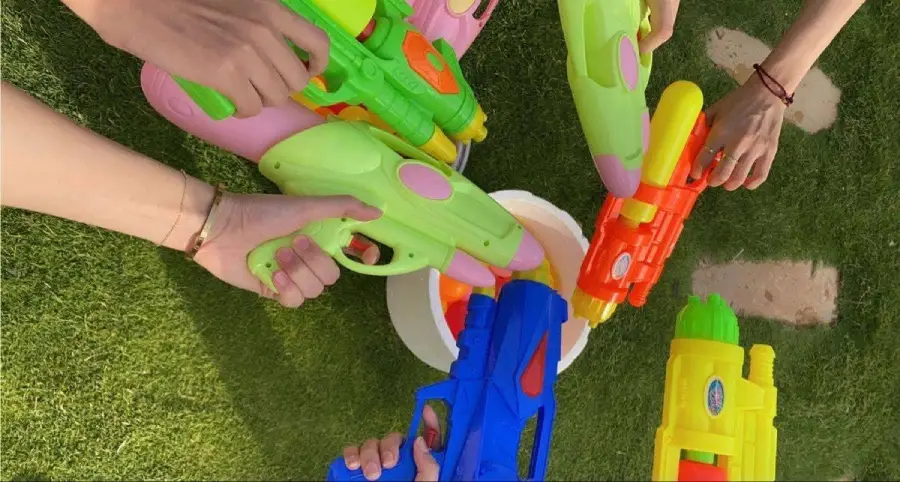 Five hands holding colorful water guns over a white bucket on green grass, preparing for a water fight.