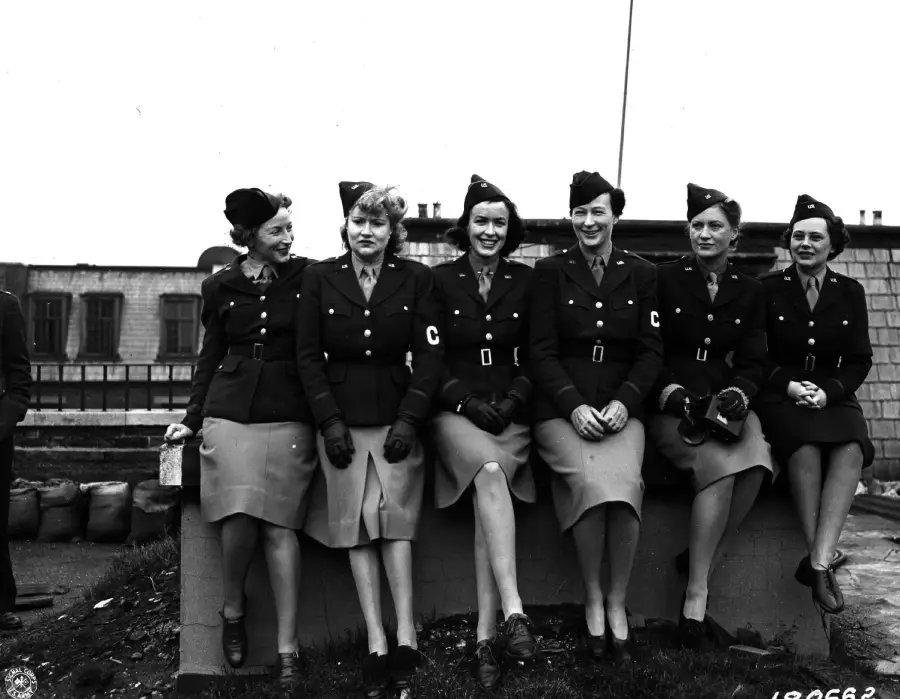 Six women in military uniforms sit on a low wall outdoors, smiling. The uniforms have skirts, double-breasted jackets, hats, and armbands with a "C". Buildings and sandbags are visible in the background.
