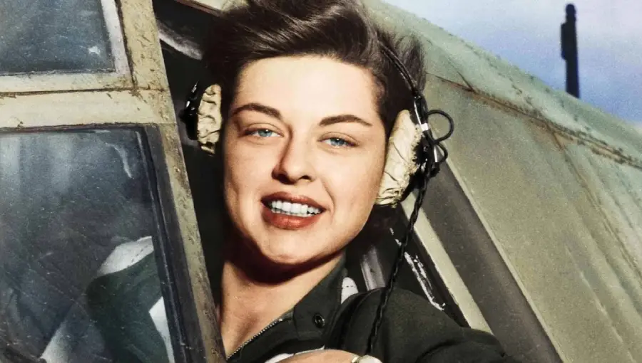 A smiling woman wearing a headset leans out of the cockpit window of an aircraft, with short dark hair and a green uniform visible.