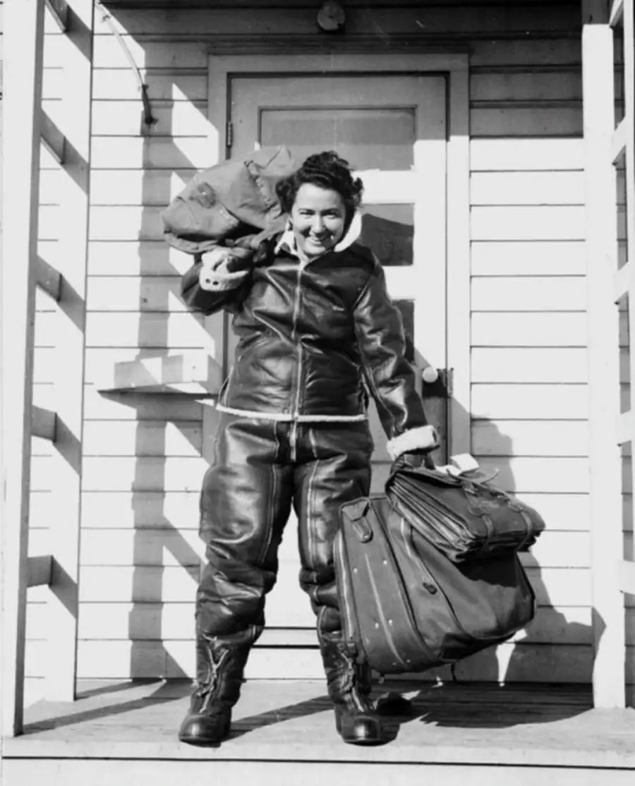 A person in a heavy leather flight suit stands on a porch, smiling, holding a large bag over one shoulder and another bag in the other hand, in front of a paneled door and white wooden wall.