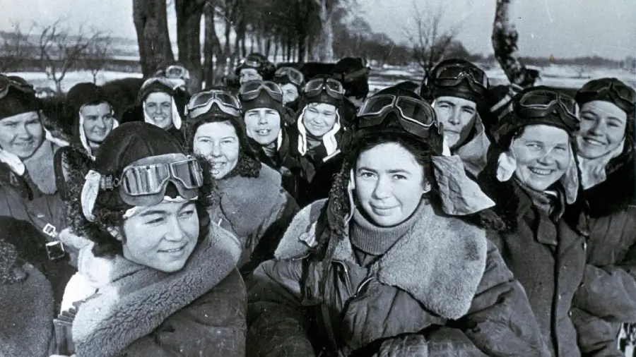 A group of women pilots in winter gear and aviator goggles stand outdoors, smiling at the camera. Snow covers the ground and bare trees line the background.