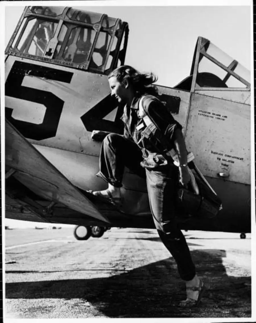 A woman in a flight suit steps out of a vintage military aircraft numbered 54, holding a helmet, with wind blowing her hair. The photo is black and white.