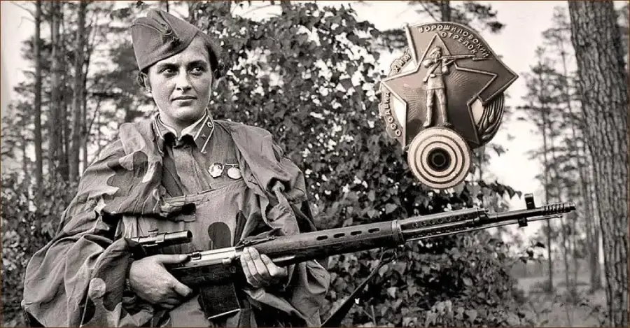 A woman in military uniform holds a rifle, standing outdoors with trees in the background. Medals are on her chest, and a large badge or medal is shown prominently to the right of her.
