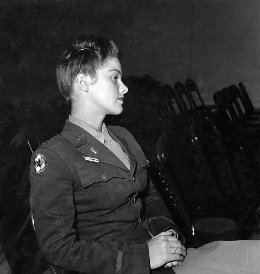 A young woman in a military uniform sits in profile, hands folded in her lap, with a row of empty chairs visible in the background.