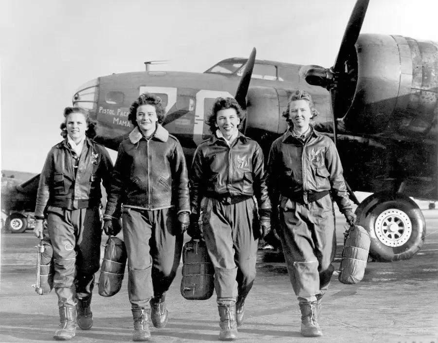 Four women pilots in flight suits stand side by side, smiling, holding helmets, in front of a large military aircraft with visible propellers. The background shows clear skies and the plane's nose art.