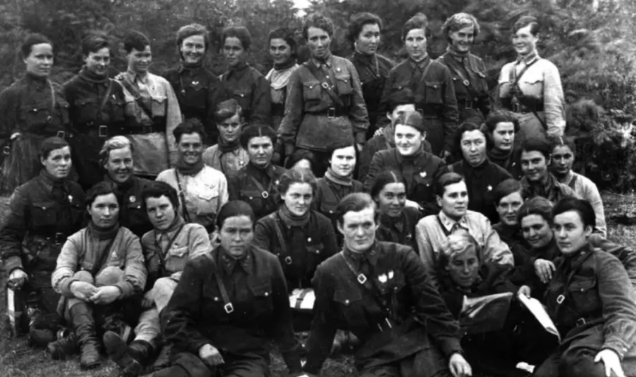 A black and white photo of a group of women in military uniforms, sitting and standing closely together outdoors, posing for the camera with neutral expressions.
