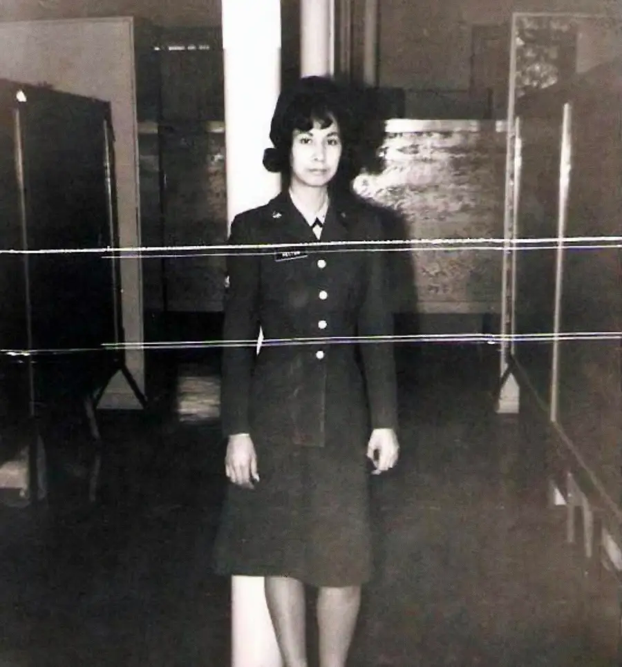 A woman in a military uniform stands indoors in front of a white column. The black and white photo has horizontal white lines running across it, showing signs of damage or distortion.