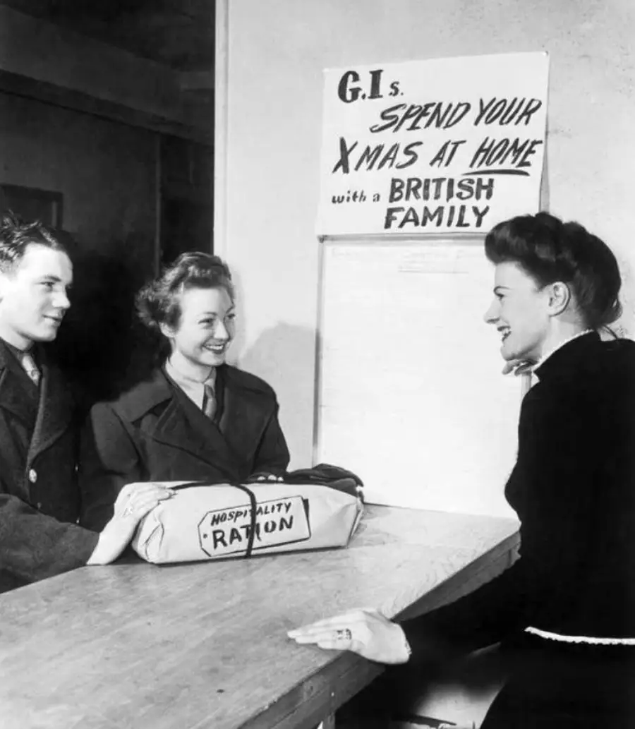 Two women and a man smile at a counter. A sign behind them says, “G.I.s, spend your Xmas at home with a British family.” A package labeled “Hospitality Ration” sits on the counter.