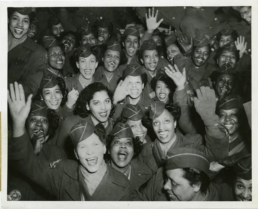 A group of smiling Black women in military uniforms wave and look at the camera in a crowded, joyful scene.