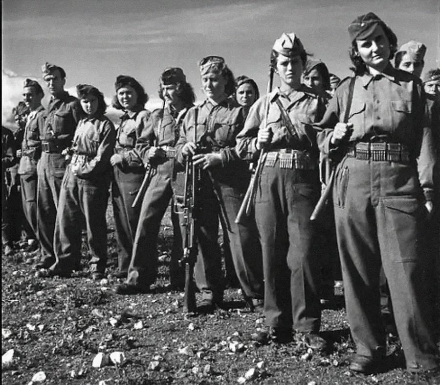 A group of women soldiers in uniform stand in a line outdoors, holding rifles and wearing military caps, with a cloudy sky in the background.