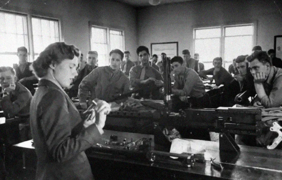 A woman stands at the front of a classroom, holding papers, while several men seated at desks watch her; the setting appears to be a mid-20th-century technical or engineering class.