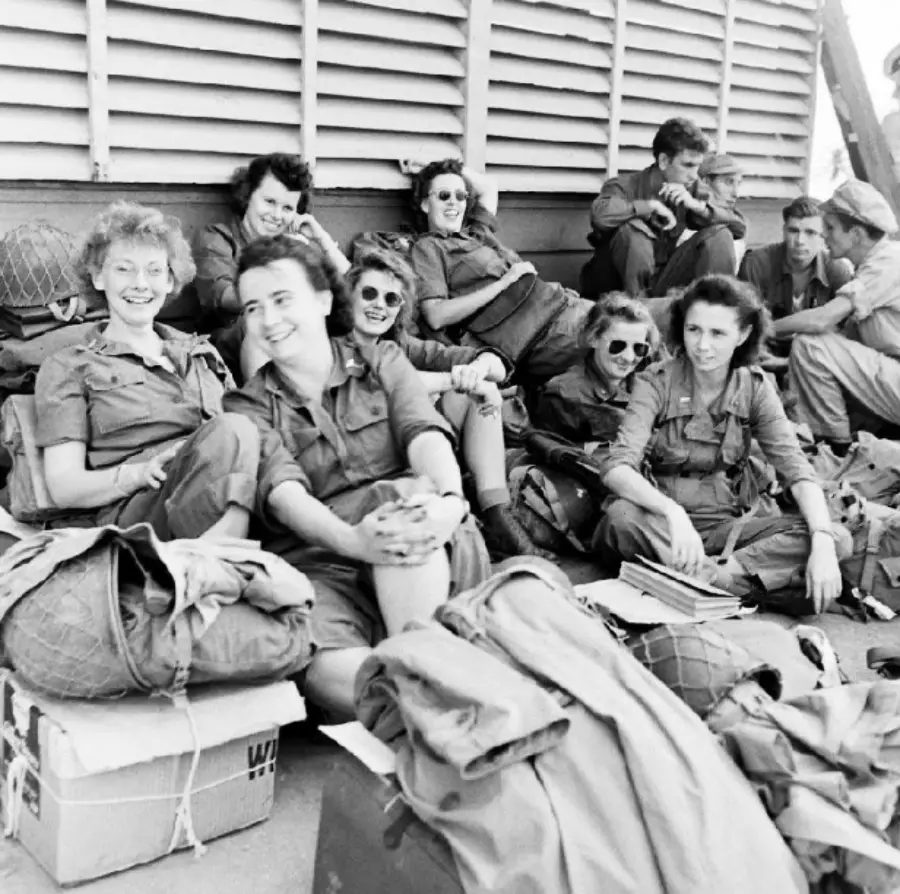 A group of women in military uniforms sit and relax together outdoors, some smiling and wearing sunglasses, surrounded by parachutes and gear.
