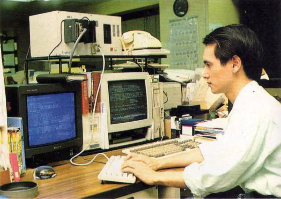 A man sits at a desk typing on a keyboard, with two CRT monitors displaying text. The desk is cluttered with office supplies, books, and equipment, including a telephone and electronics. The setting appears to be an office.
