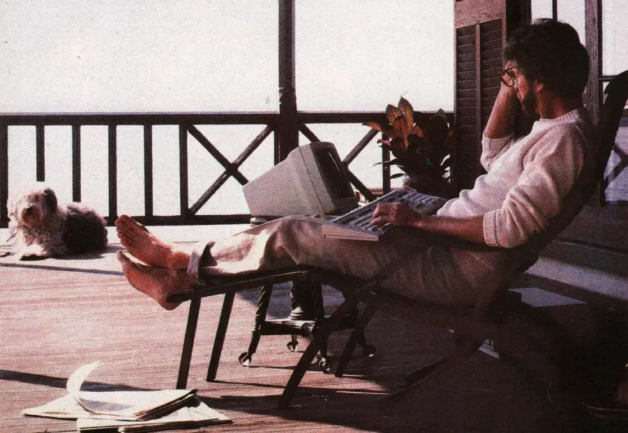 A person sits on a lounge chair on a wooden porch, using a vintage computer on their lap. Papers are scattered on the floor, and a dog lies in the background near the porch railing.